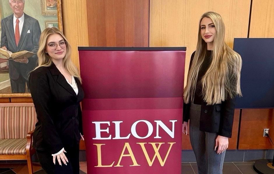 Two young women wearing professional business suits smile while standing on either side of a maroon sign that reads "ELON LAW."