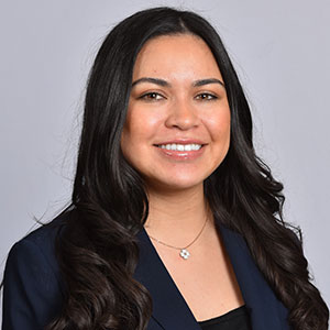 Professional headshot of Alisson Rodriguez, a smiling woman with long, wavy dark hair, wearing a navy blue blazer and a delicate necklace against a plain light gray background.