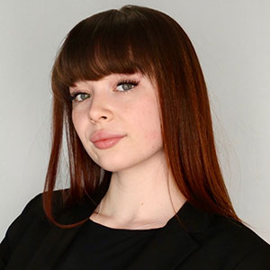 Professional headshot of a young woman with long, straight, reddish-brown hair and straight-across bangs, wearing a black blazer and smiling subtly against a light gray background.