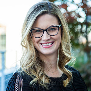 Professional outdoor headshot of Chelsey Glasson, a smiling woman with shoulder-length wavy blonde hair and black glasses, wearing a black textured top.