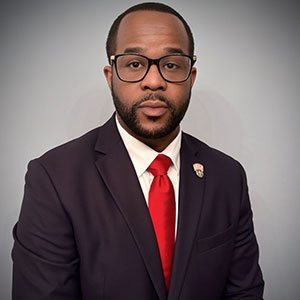 Professional headshot of Dwight White, a Black man with a trimmed beard and black-framed glasses, wearing a dark navy suit jacket, white collared shirt, and a bright red tie against a gray background.