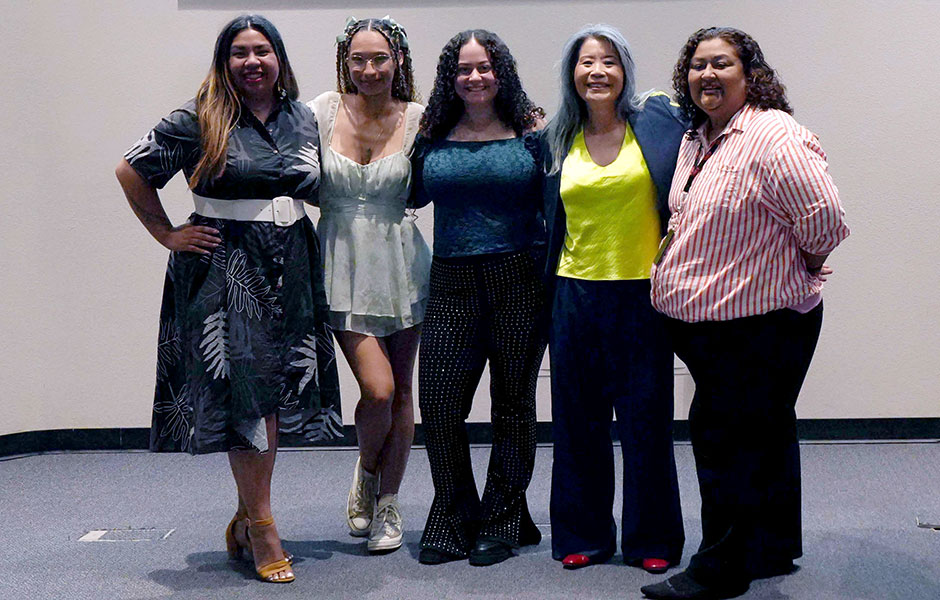 A medium full shot of five smiling women standing arm-in-arm in a row against a plain beige wall. From left to right: the first woman wears a dark floral print dress with a wide white belt; the second wears a light-colored, short, sleeveless dress and sneakers; the third wears a dark off-the-shoulder top and sparkly black trousers; the fourth wears a bright yellow top under a dark jacket with wide-leg pants; and the fifth wears a red and white striped button-down shirt with black pants.