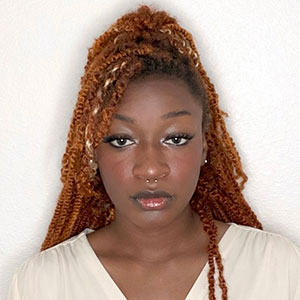 Professional headshot of Noreyahnna Black, a young Black woman with long, copper-colored textured twists styled half-up. She is wearing a cream v-neck top and a small gold septum piercing against a plain white background.