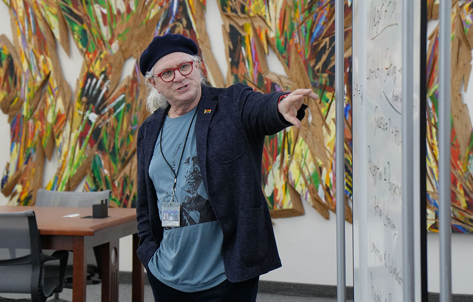 Professor Patrick Chapin gesturing toward a whiteboard with handwritten notes during a presentation, wearing a blue beret, glasses, a dark blazer, and a graphic t-shirt, set against a large, colorful abstract art piece on the wall behind him.
