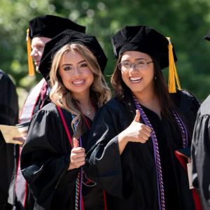 Two feamle students in graduation regalia smile and give thumbs up to the camera