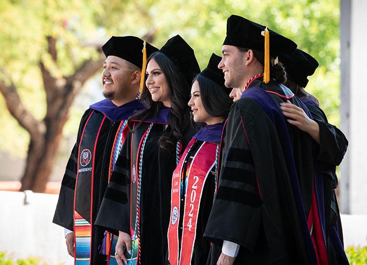 five graduates stand together in cap and gown smiling at the camera