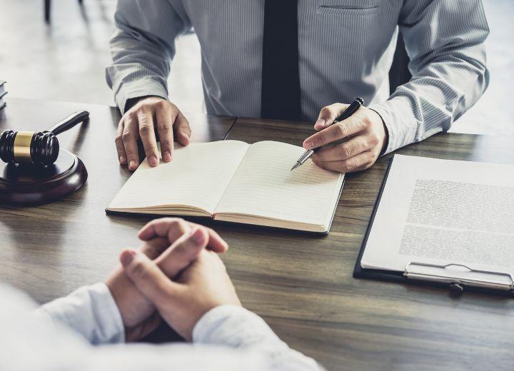 two people sit at desk with a gavel, open book, pen, and clipboard laying on the desk